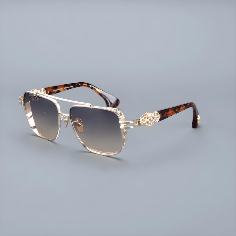 May include: Gold-coloured sunglasses with gradient lenses. The sunglasses have a double bridge and decorative detailing on the temples. The arms feature a tortoiseshell pattern. The lenses transition from light to dark.
