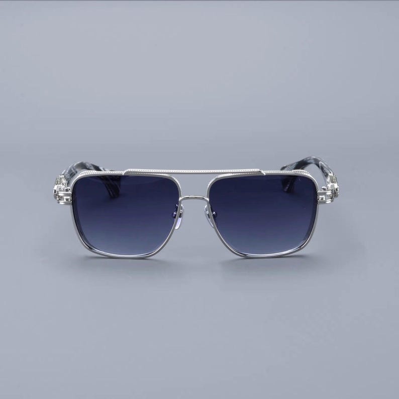 May include: Silver-framed sunglasses with a double bridge and gradient blue lenses. The temples feature a patterned design. The sunglasses have a square shape and a modern aesthetic.