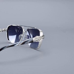 May include: Silver-framed sunglasses with square lenses and a double bridge. The temples have a marbled pattern and are adorned with decorative embellishments. The lenses are a dark blue colour, offering a stylish look.