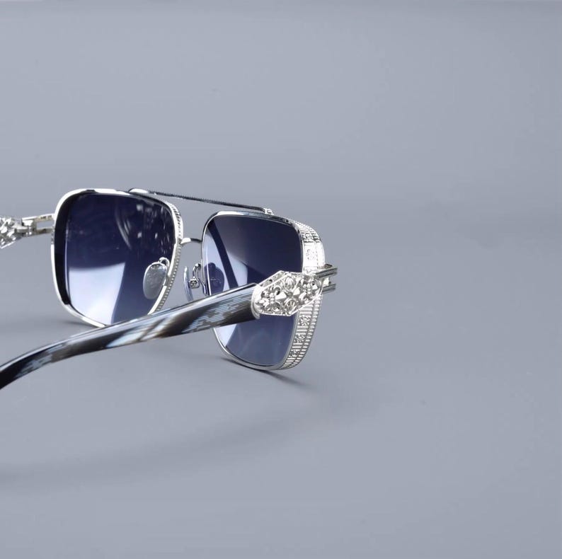 May include: Silver-framed sunglasses with square lenses and a double bridge. The temples have a marbled pattern and are adorned with decorative embellishments. The lenses are a dark blue colour, offering a stylish look.