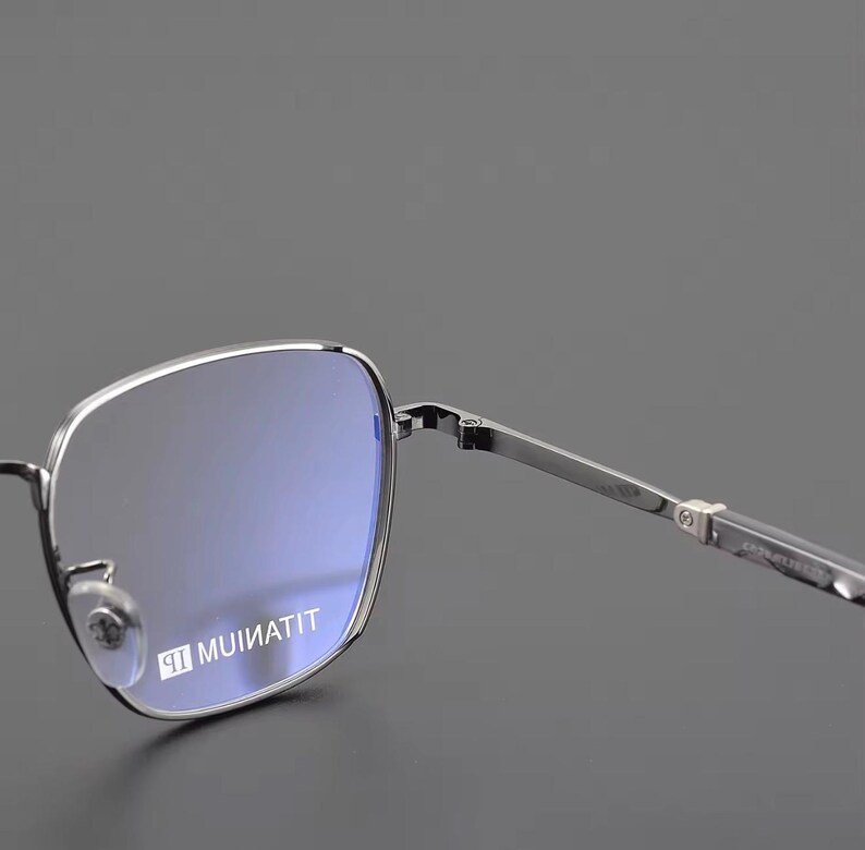 Puede incluir: Gafas con montura plateada y lentes cuadradas. Las lentes tienen un tinte azul y la montura es de color plateado brillante. El texto "MUINATIT" est&aacute; impreso en la lente. Dise&ntilde;o moderno y minimalista.