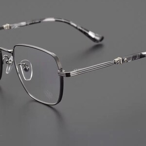 Puede incluir: Primer plano de gafas con montura negra y plateada. Las lentes son transparentes y las patillas tienen un dise&ntilde;o veteado en blanco y negro. La montura tiene forma cuadrada con un dise&ntilde;o decorativo en las bisagras.
