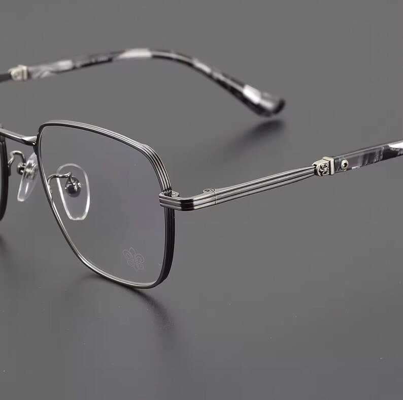 Puede incluir: Primer plano de gafas con montura negra y plateada. Las lentes son transparentes y las patillas tienen un dise&ntilde;o veteado en blanco y negro. La montura tiene forma cuadrada con un dise&ntilde;o decorativo en las bisagras.