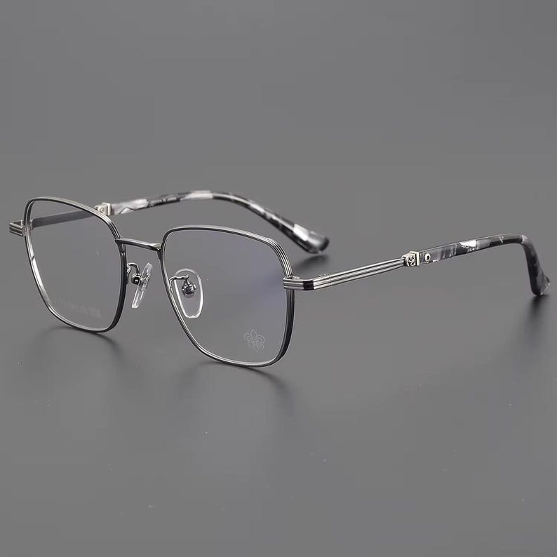 Puede incluir: Gafas con montura de metal gris oscuro y lentes transparentes. Las patillas presentan un patr&oacute;n veteado en blanco y negro. Las gafas tienen una forma geom&eacute;trica y un peque&ntilde;o logotipo en la lente inferior derecha.
