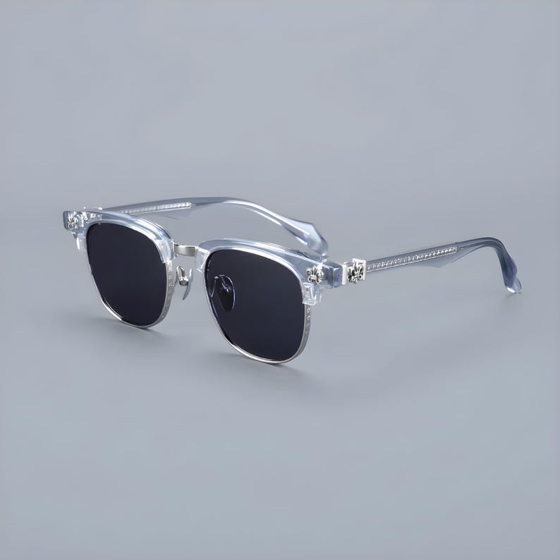 May include: Clear-framed sunglasses with dark lenses. The sunglasses have a square shape with silver metal accents and decorative embellishments on the temples. The arms are a clear, light blue color.