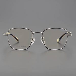 Puede incluir: Gafas de montura cuadrada con montura de metal plateado y detalles en negro. Las lentes son tintadas y las patillas presentan un dise&ntilde;o estampado. El texto "TITANIUM TE" es visible en la montura.