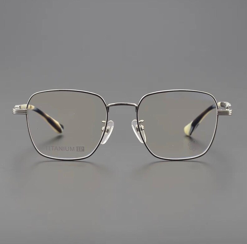 Puede incluir: Gafas de montura cuadrada con montura de metal plateado y detalles en negro. Las lentes son tintadas y las patillas presentan un dise&ntilde;o estampado. El texto "TITANIUM TE" es visible en la montura.
