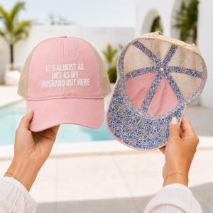 May include: A pink trucker hat with a tan mesh back and the white text IT'S ALMOST AS HOT AS MY HUSBAND OUT HERE on the front. The interior of the hat features a blue, pink, and white floral pattern lining along the seams and the brim edge.
