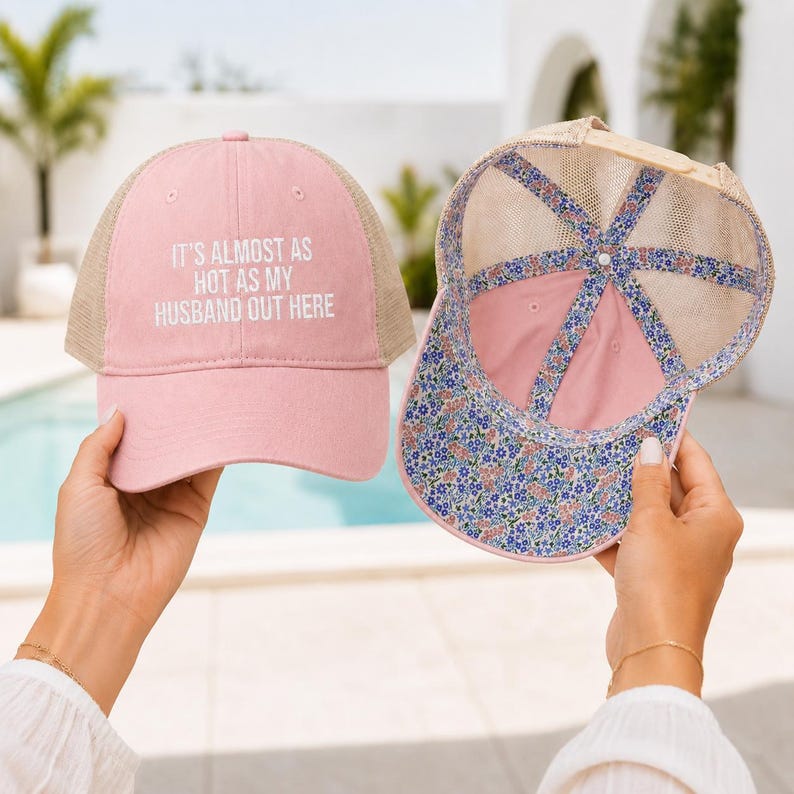 May include: A pink trucker hat with a tan mesh back and the white text IT'S ALMOST AS HOT AS MY HUSBAND OUT HERE on the front. The interior of the hat features a blue, pink, and white floral pattern lining along the seams and the brim edge.