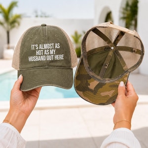 May include: An olive green trucker hat with a mesh back and the white text IT'S ALMOST AS HOT AS MY HUSBAND OUT HERE printed on the front. The interior of the hat features a camouflage pattern on the brim and inner panels with a mesh structure.