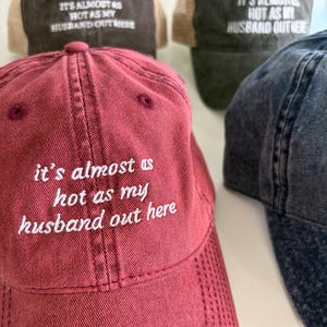 May include: A collection of baseball caps in maroon, dark brown, and charcoal gray, each featuring the embroidered white text 'it's almost as hot as my husband out here' on the front panel. The caps have a distressed finish and structured crowns.