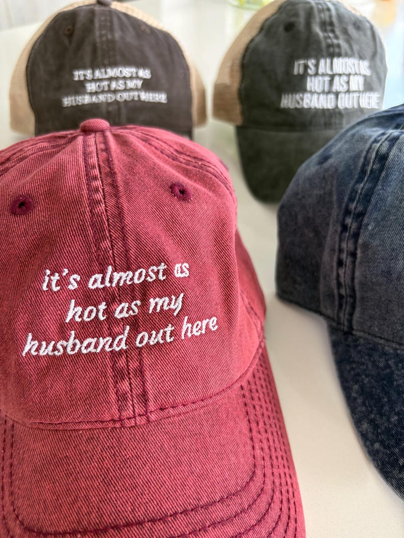 May include: A collection of baseball caps in maroon, dark brown, and charcoal gray, each featuring the embroidered white text 'it's almost as hot as my husband out here' on the front panel. The caps have a distressed finish and structured crowns.