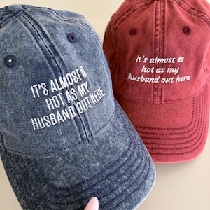 May include: Two baseball caps, one in acid-wash blue and one in acid-wash red, both featuring white embroidered text that reads: IT'S ALMOST AS HOT AS MY HUSBAND OUT HERE. Each cap has a six-panel construction with a button on top and eyelets.