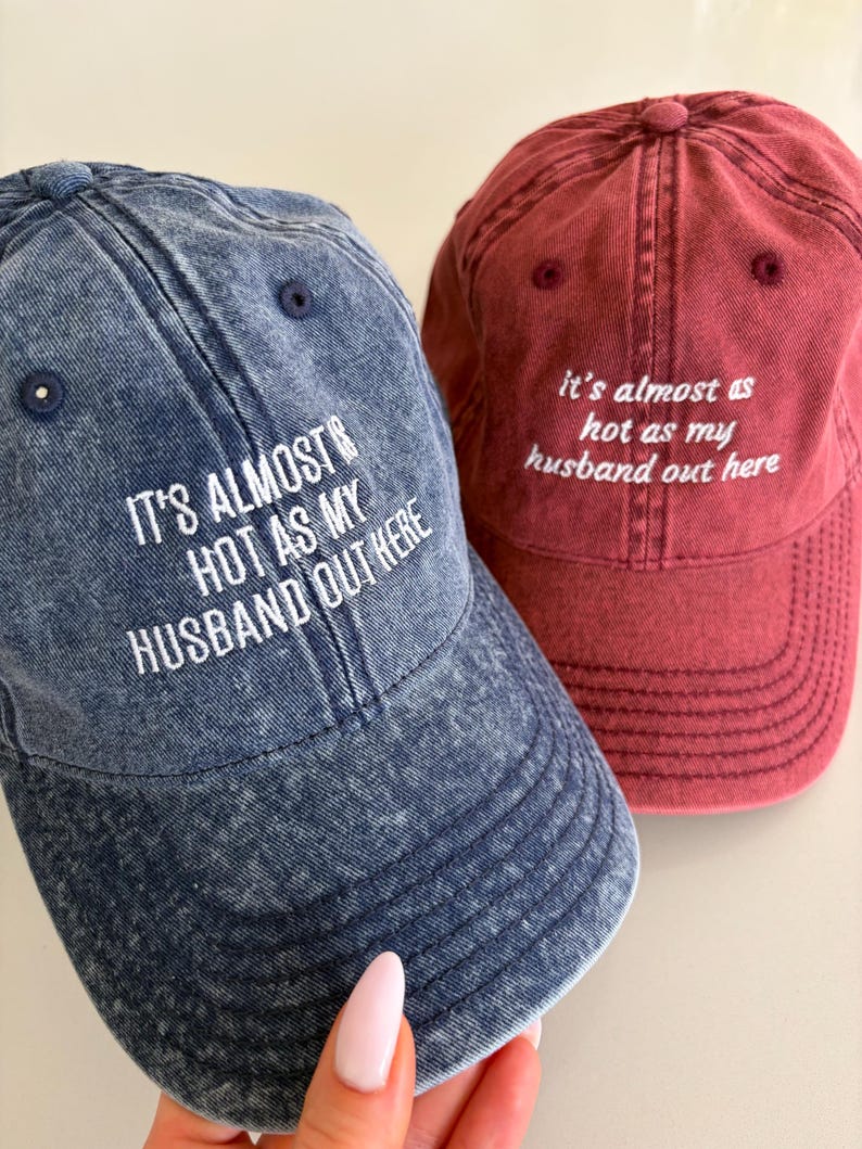 May include: Two baseball caps, one in acid-wash blue and one in acid-wash red, both featuring white embroidered text that reads: IT'S ALMOST AS HOT AS MY HUSBAND OUT HERE. Each cap has a six-panel construction with a button on top and eyelets.