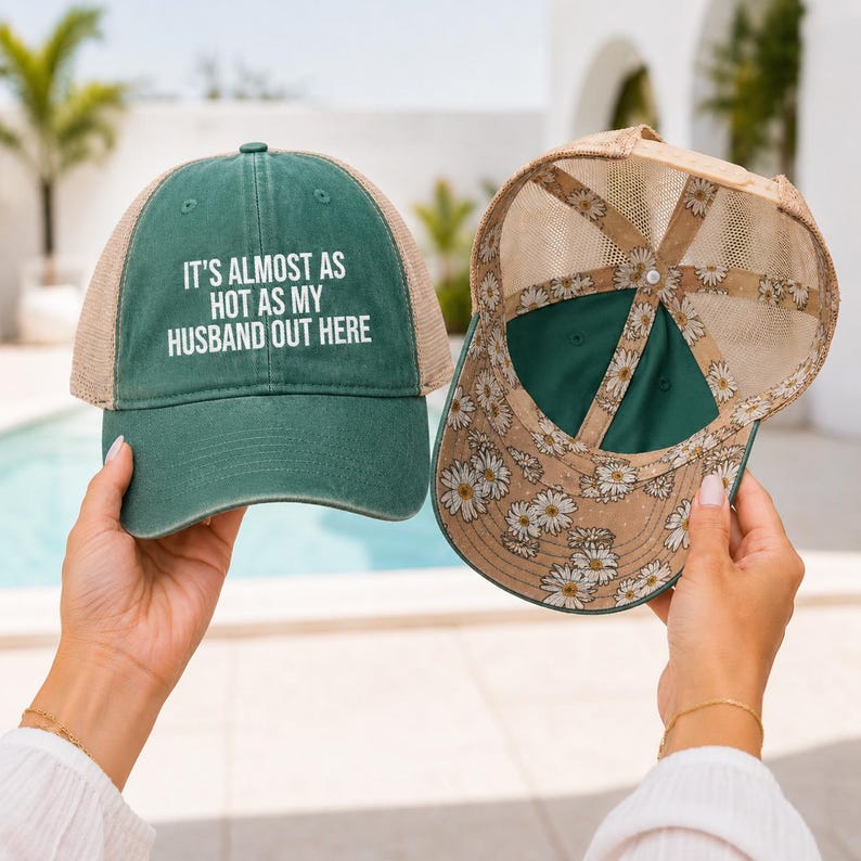 May include: A dark green baseball cap with a tan mesh back and the white text IT'S ALMOST AS HOT AS MY HUSBAND OUT HERE on the front. A second cap is shown from the inside, featuring a tan mesh lining with a white and yellow daisy floral pattern.