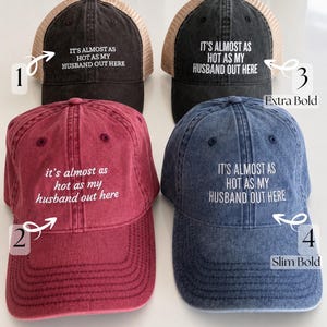 May include: Four trucker hats in black, red, and blue with mesh backs. Each hat features the white embroidered text 'IT'S ALMOST AS HOT AS MY HUSBAND OUT HERE' in varying font styles labeled 1 through 4, categorized as Classic Fonts and Bold Fonts.