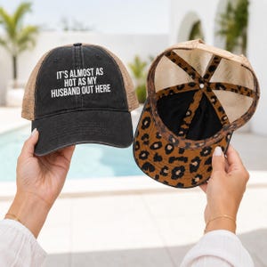 May include: A black baseball cap with a tan mesh back and the white text IT'S ALMOST AS HOT AS MY HUSBAND OUT HERE on the front, paired with a second cap featuring a tan mesh back and a brown and black leopard print pattern on the underside of the brim.