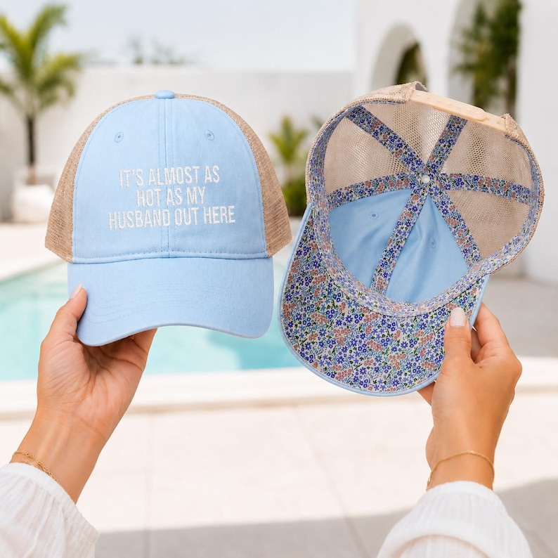May include: A light blue baseball cap with a tan mesh back and the white embroidered text IT'S ALMOST AS HOT AS MY HUSBAND OUT HERE. A second cap is shown from the inside, featuring a blue, pink, and green floral pattern on the inner lining.