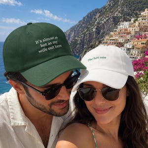 May include: A dark green baseball cap with the white embroidered text 'It's almost as hot as my wife out here' and a white baseball cap with the green embroidered text 'The hot one', both featuring a curved brim and a structured crown design.