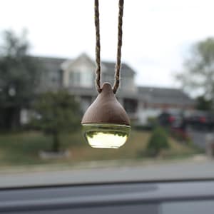 May include: A car air freshener with a wooden top and a glass bottom filled with a yellow liquid. The air freshener is suspended by a brown and gold rope, hanging in front of a blurred outdoor scene.