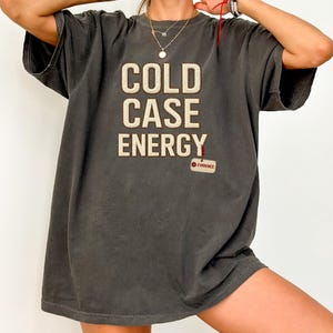 Puede incluir: Camiseta gris oscuro con la frase "COLD CASE ENERGY" en letras texturizadas color crema. Una pequeña etiqueta con "EVIDENCE" está adjunta. La camiseta tiene mangas cortas y un corte holgado.