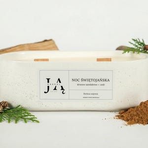 May include: A rectangular, speckled white candle with three wooden wicks. The candle is labeled "Noc Świętojańska" and is surrounded by wood, greenery, and a pile of brown powder. The candle is a home decor item.