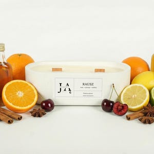 Puede incluir: Una vela blanca ovalada con mechas de madera, rodeada de fruta fresca, especias y pequeñas botellas de líquido. Naranjas, limones, limas y cerezas están dispuestas alrededor de la vela, creando una exhibición vibrante y aromática.