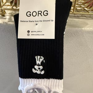 Può includere: Calzini neri con polsino bianco a balze e grafica a teschio. I calzini hanno la scritta "GORG" stampata su un'etichetta, insieme alla frase "Balance Starts from the Ground Up" e alle informazioni sui social media.