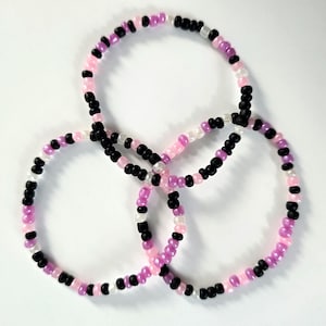 May include: Three beaded bracelets in a circular design. The bracelets are made of small beads in black, pink, white, and purple. The bracelets are intertwined, creating a unique pattern.