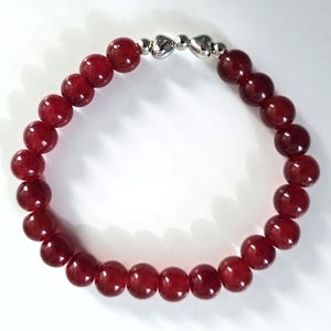 Könnte beinhalten: Ein rotes Perlenarmband mit einem silbernen Verschluss. Das Armband besteht aus runden, polierten roten Perlen, die aufgefädelt sind. Der Verschluss besteht aus kleinen, herzförmigen silbernen Gliedern.