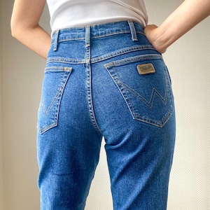 May include: Blue denim Wrangler jeans with a high waist and a classic five-pocket design. The jeans feature a gold-colored Wrangler patch on the back pocket and a signature stitching detail. The jeans are paired with a white t-shirt.
