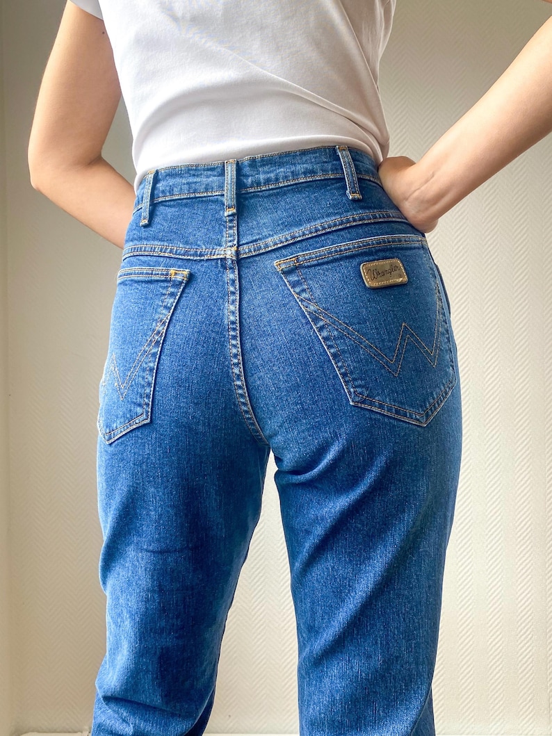 May include: Blue denim Wrangler jeans with a high waist and a classic five-pocket design. The jeans feature a gold-colored Wrangler patch on the back pocket and a signature stitching detail. The jeans are paired with a white t-shirt.
