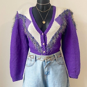 Peut inclure: Un cardigan en tricot violet avec un motif en losanges sur les épaules et la poitrine, associé à un short en jean délavé. Le cardigan a des boutons dorés et des franges. Une ceinture chaîne dorée et des colliers superposés complètent la tenue.
