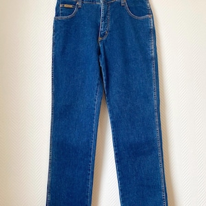 May include: Classic blue denim jeans with a straight leg cut. The jeans feature a standard five-pocket design, a button and zipper closure, and a small logo patch on the front pocket. The jeans are hanging on a metal hanger.