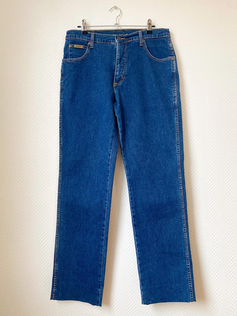 May include: Classic blue denim jeans with a straight leg cut. The jeans feature a standard five-pocket design, a button and zipper closure, and a small logo patch on the front pocket. The jeans are hanging on a metal hanger.