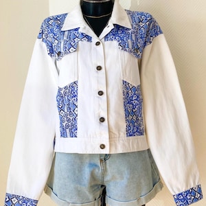 Peut inclure: Une veste en jean blanche avec des empiècements à motifs bleus et blancs sur les épaules, les poches de poitrine et les poignets. La veste présente un col classique, des manches longues et une fermeture à boutons métalliques, avec un short en jean.