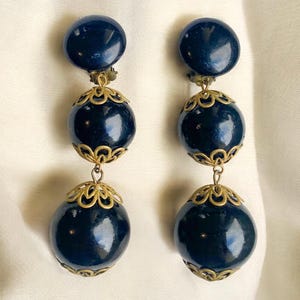 Peut inclure: Une paire de boucles d'oreilles clip vintage bleu marine. Chaque boucle d'oreille comporte trois sphères bleu marine reliées par des filigranes dorés ornés. La sphère supérieure est plus petite, les deux sphères inférieures augmentant en taille.