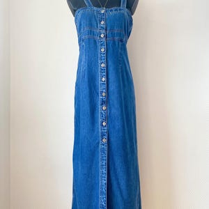 Peut inclure: Une robe longue en jean bleu dotée d'une encolure carrée, de larges bretelles et d'une patte de boutonnage frontale sur toute la longueur avec des boutons métalliques. Le vêtement présente des coutures apparentes sur la poitrine.