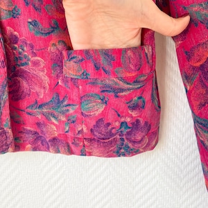 May include: A bright pink corduroy jacket featuring a dark purple and teal floral pattern. The jacket includes a visible front patch pocket with a horizontal opening, showcasing the textured ribbed fabric and the intricate botanical print design.