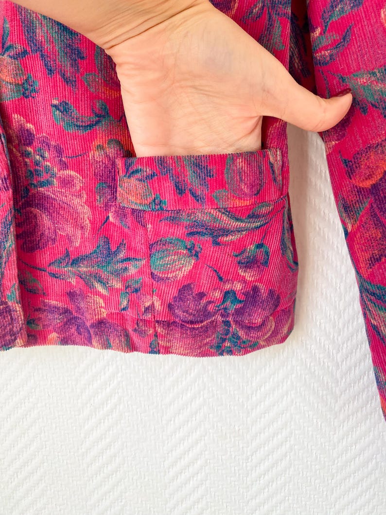 May include: A bright pink corduroy jacket featuring a dark purple and teal floral pattern. The jacket includes a visible front patch pocket with a horizontal opening, showcasing the textured ribbed fabric and the intricate botanical print design.
