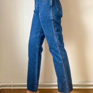 May include: Blue denim jeans with a straight leg cut. The jeans have a classic five-pocket design and a medium wash. The model is wearing gold strappy heels. The jeans are a versatile wardrobe staple.
