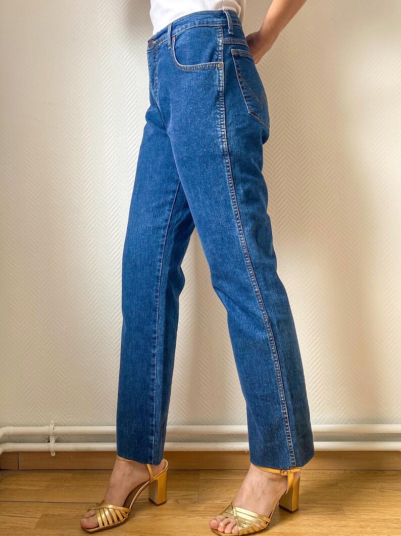 May include: Blue denim jeans with a straight leg cut. The jeans have a classic five-pocket design and a medium wash. The model is wearing gold strappy heels. The jeans are a versatile wardrobe staple.