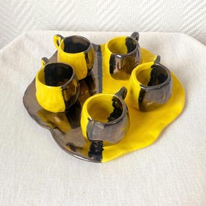Peut inclure: Un service à expresso en céramique avec un motif jaune et noir. L'ensemble comprend cinq petites tasses et un plateau assorti. Les tasses et le plateau ont une finition brillante. Les tasses mesurent environ 5 cm de haut.