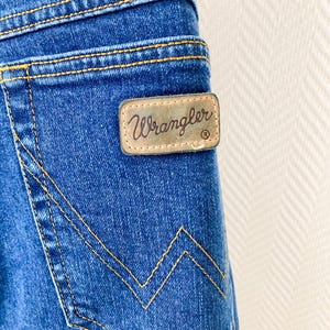 May include: Close-up of blue denim jeans with yellow stitching. A brown leather patch with the brand name "Wrangler" is visible on the back pocket. The jeans have a classic five-pocket design.