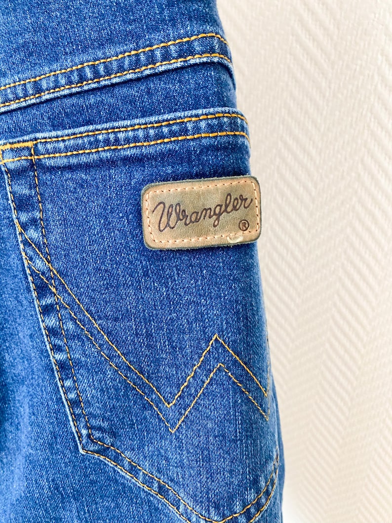 May include: Close-up of blue denim jeans with yellow stitching. A brown leather patch with the brand name "Wrangler" is visible on the back pocket. The jeans have a classic five-pocket design.