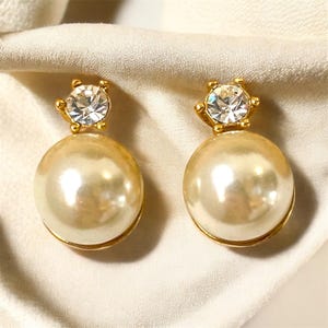 Peut inclure: Une paire de boucles d'oreilles dorées. Chaque boucle d'oreille est composée d'une grosse perle ronde de couleur crème, surmontée d'une petite pierre précieuse transparente et étincelante sertie dans une couronne dorée. Sur tissu crème.
