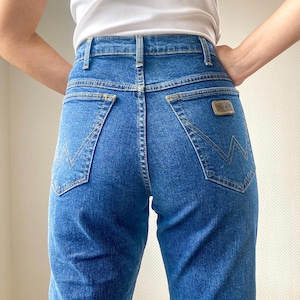 May include: Blue denim jeans with a high waist and a classic five-pocket design. The jeans feature a straight leg cut and a small, rectangular patch on the back pocket. The stitching is a contrasting gold color.