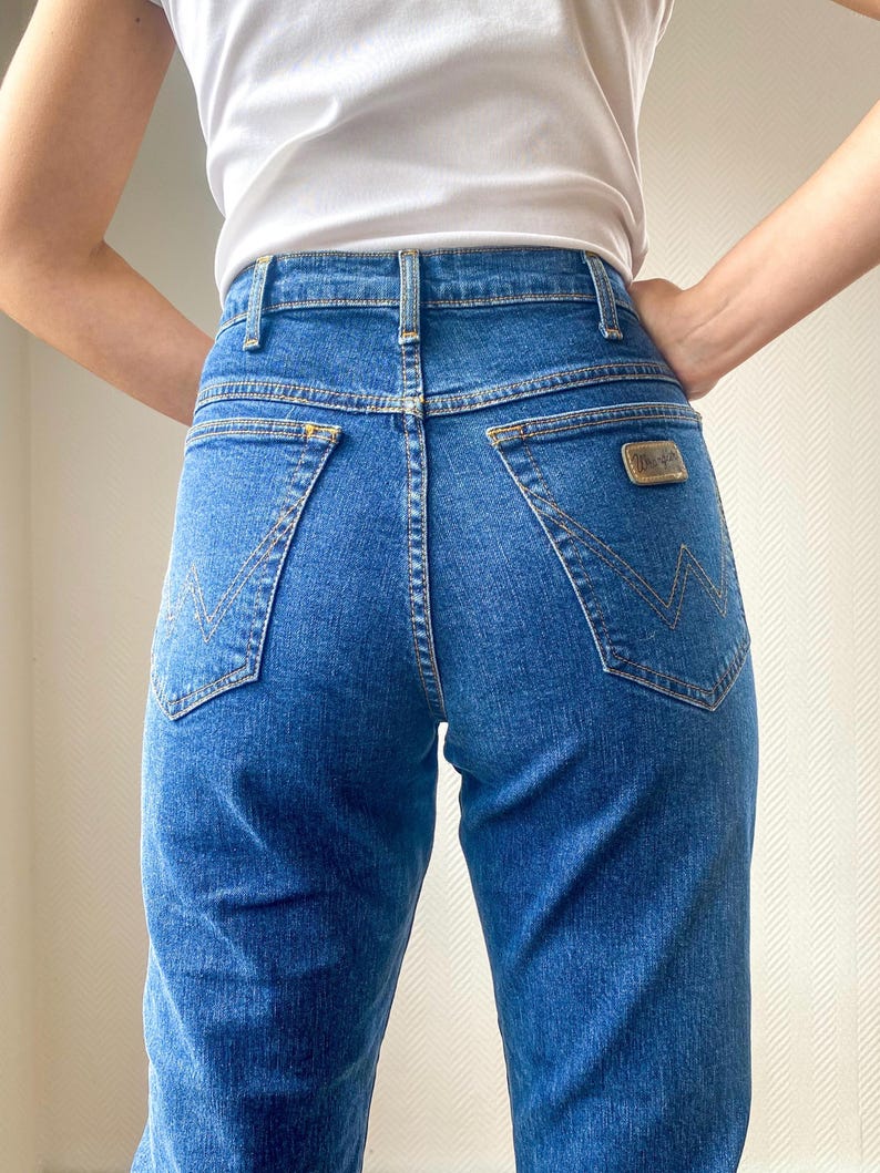 May include: Blue denim jeans with a high waist and a classic five-pocket design. The jeans feature a straight leg cut and a small, rectangular patch on the back pocket. The stitching is a contrasting gold color.