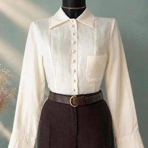 May include: A long-sleeved cream-colored button-down shirt featuring a pointed collar, a single chest pocket with subtle embroidery, and a row of small buttons. The shirt is tucked into dark brown trousers cinched with a brown leather belt.