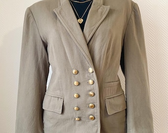 May include: A tan double-breasted blazer with gold-colored buttons. The blazer has two front pockets and a notched lapel. The jacket is displayed on a black mannequin with a gold necklace. The blazer is paired with denim shorts.